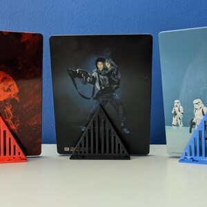 May include: Three triangular stands made of plastic in red, black, and blue, each holding a different Star Wars movie poster. The posters are all in the same style, with a dark background and a single image of a character or scene.