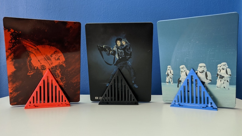 May include: Three triangular stands made of plastic in red, black, and blue, each holding a different Star Wars movie poster. The posters are all in the same style, with a dark background and a single image of a character or scene.