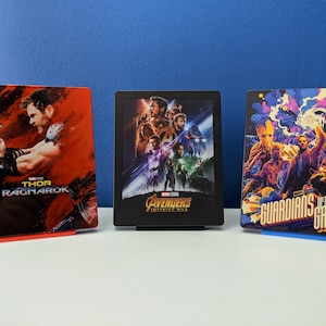 May include: Three Marvel movie Blu-ray cases with colorful artwork. The left case features Thor Ragnarok with Thor holding a sword. The middle case features Avengers Infinity War with a group of superheroes. The right case features Guardians of the Galaxy with a group of characters in a colorful nebula.