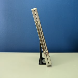 May include: Two silver-colored metal cases are stacked on top of each other. The cases are standing upright on a black plastic stand. The cases are on a white surface in front of a blue wall.