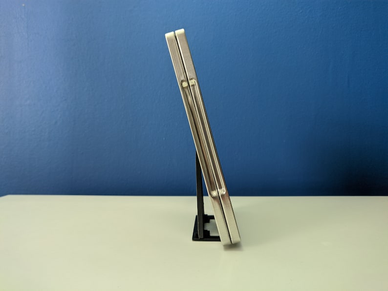 May include: Two silver-colored metal cases are stacked on top of each other. The cases are standing upright on a black plastic stand. The cases are on a white surface in front of a blue wall.