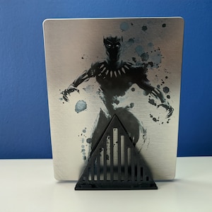 May include: A black and white image of Black Panther on a silver metal case. The case is standing on a black triangular stand with vertical bars.
