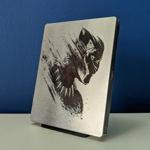 May include: A silver steel case with a black and white image of Black Panther, the superhero from the Marvel Cinematic Universe. The case is for a Blu-ray or DVD disc.