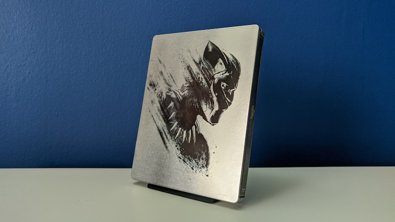 May include: A silver steel case with a black and white image of Black Panther, the superhero from the Marvel Cinematic Universe. The case is for a Blu-ray or DVD disc.