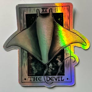 Könnte beinhalten: Holografischer Aufkleber mit einem Mantarochen-Design in einem Tarotkartenrahmen. Die Karte trägt die Aufschrift "The Devil" mit der Nummer "XV" oben. Der Aufkleber hat einen regenbogenfarbenen, irisierenden Effekt.