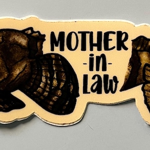 May include: A sticker with a fish illustration and the words "MOTHER-IN-LAW" in black. The fish is brown and black with detailed scales. The sticker has a cream-colored border and the words "scaled up" are in the bottom right corner.