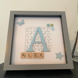 Könnte beinhalten: Ein gerahmtes Kinderzimmer-Dekor mit einem hellblauen Buchstaben A, einem Schmetterling, einem Stern und dem Namen "ALEX", der mit Scrabble-Steinen ausgeschrieben ist.