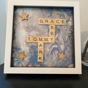 May include: A framed artwork with a white border. The artwork features wooden letter tiles spelling out names against a blue and white marbled background. Wooden star accents are placed around the names.