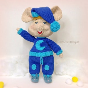 Puede incluir: Un juguete de peluche de ratón tejido a crochet que lleva un pijama azul con un diseño de media luna y un gorro de dormir azul. El juguete tiene un cuerpo marrón claro y una nariz azul claro.