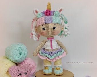 Pode incluir: Uma boneca de unicórnio de crochê com um vestido rosa, roxo e branco, cabelo de lã branco e um coração rosa na frente de novelos de lã.