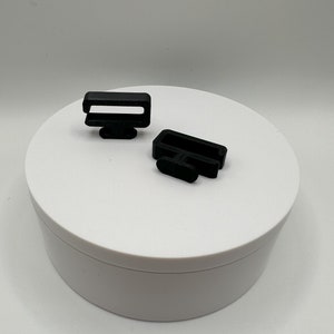May include: Two small, black plastic clips are displayed on a white surface. Each clip has a rectangular loop and a smaller, rectangular base. The clips are likely used for securing straps or other items.