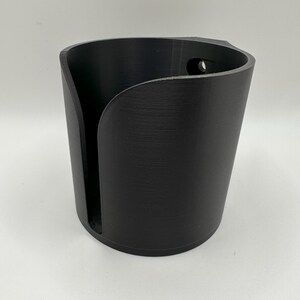 May include: A black, cylindrical cup holder with a curved opening on one side. The holder has a textured surface and a small hole near the top. The design is simple and modern, suitable for various settings.