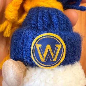 May include: A blue knit hat with a yellow scarf and a white pom-pom. The hat features a yellow and blue circular emblem with a large yellow "W" in the center. The hat is made of a soft, knitted material.