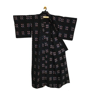 May include: Black kimono with a repeating pattern of small pink and blue squares. The kimono is hanging on a black hanger against a white background. The sleeves are wide and the garment is open in the front.