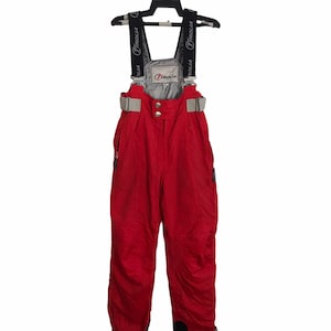 May include: Red ski bibs with black and grey accents, hanging on a black hanger. The bibs have adjustable straps with the brand name "Tyrolia" printed on them. The bibs have a grey panel at the chest and waist with the brand logo.