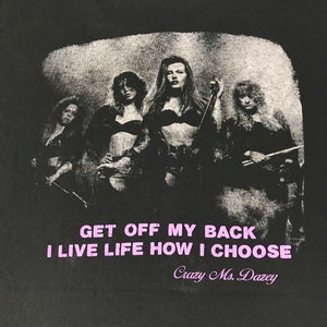 Puede incluir: Camiseta negra con una imagen en escala de grises de cuatro mujeres con instrumentos musicales. El texto rosa dice "GET OFF MY BACK I LIVE LIFE HOW I CHOOSE", con "Crazy Ms. Dazey" debajo.