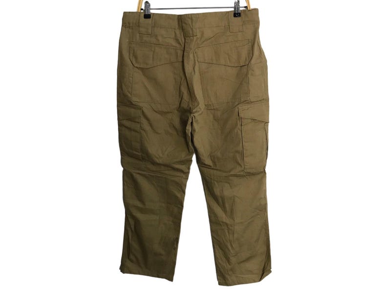 Pu&ograve; includere: Pantaloni cargo color kaki appesi a una gruccia di legno. I pantaloni hanno pi&ugrave; tasche sui lati e sul retro. Il tessuto sembra essere un materiale resistente e di peso medio.