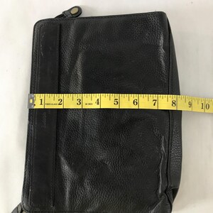 May include: Black leather wallet with a textured surface. The wallet is rectangular and measures approximately 23 cm in length. It features a zipper closure and a strap with a metal clasp.