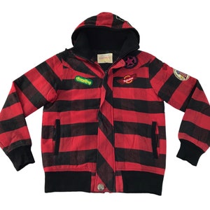 Könnte beinhalten: Rot-schwarz gestreifte Kapuzenjacke mit mehreren Aufnähern. Die Jacke hat eine schwarze Kapuze, Bündchen und Saum. Sie hat einen Reißverschluss und zwei Vordertaschen. Zu den Aufnähern gehören ein Stern, eine Flagge und Text.