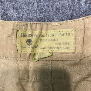 Pu&ograve; includere: Primo piano di pantaloni tattici Emerson color beige. L'etichetta recita "EMERSON Tactical Pants" con il numero articolo EM7035 e un logo a teschio. &Egrave; visibile anche il sito web www.emersongear.com. I pantaloni hanno una trama strutturata.