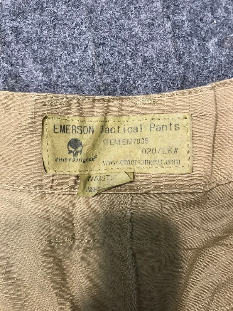 Pu&ograve; includere: Primo piano di pantaloni tattici Emerson color beige. L'etichetta recita "EMERSON Tactical Pants" con il numero articolo EM7035 e un logo a teschio. &Egrave; visibile anche il sito web www.emersongear.com. I pantaloni hanno una trama strutturata.