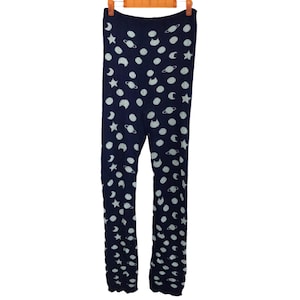 Può includere: Leggings blu navy con un motivo ripetuto di corpi celesti bianchi, tra cui pianeti, stelle e lune crescenti. I leggings sono appesi a una gruccia di legno. Il tessuto sembra morbido ed elastico.