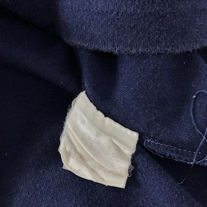 May include: Close-up of navy blue fabric with a white tag attached. The tag appears to have text printed on it. The fabric is textured and appears to be a piece of clothing.