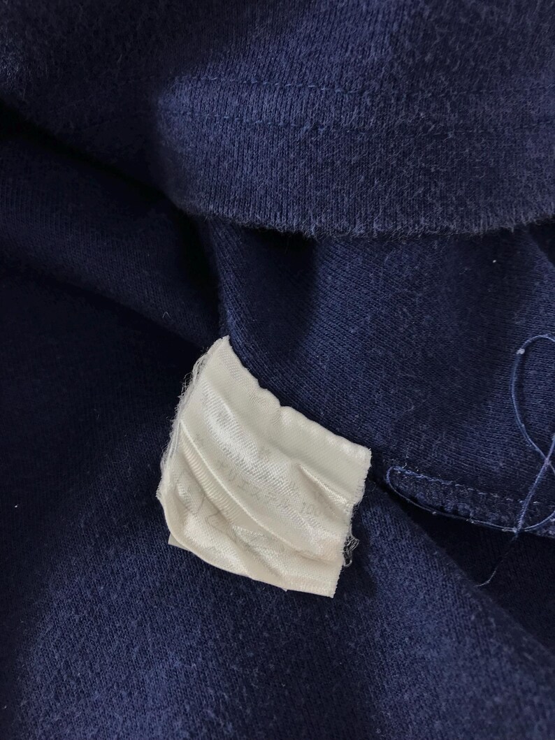 May include: Close-up of navy blue fabric with a white tag attached. The tag appears to have text printed on it. The fabric is textured and appears to be a piece of clothing.