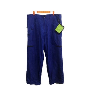 Puede incluir: Pantalones de nieve azul real con cremalleras y bolsillos negros, colgados de una percha de madera. Una pequeña bolsa verde y negra está adjunta. Los pantalones están hechos de un material resistente al agua.