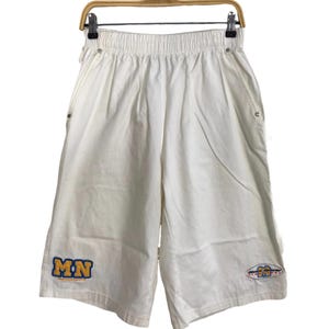 Può includere: Pantaloncini sportivi bianchi con elastico in vita e tasche laterali. I pantaloncini presentano le lettere "MN" in blu e oro sulla gamba sinistra e un logo colorato sulla gamba destra. I pantaloncini sono appesi a una gruccia di legno.