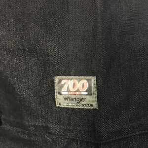 May include: Close-up of dark denim fabric with a rectangular label. The label reads "700 Series Wrangler Greensboro N.C. U.S.A." in white and red lettering on a green background. The denim appears to be a deep indigo colour.