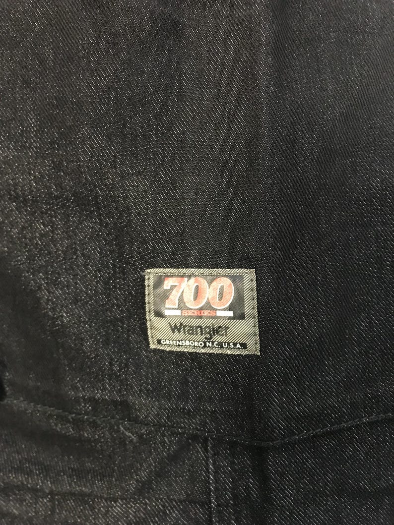May include: Close-up of dark denim fabric with a rectangular label. The label reads "700 Series Wrangler Greensboro N.C. U.S.A." in white and red lettering on a green background. The denim appears to be a deep indigo colour.