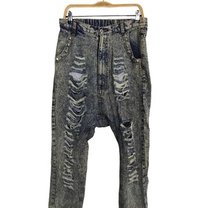 Op de afbeelding: Acid wash denim jeans met distressed details, hangend aan een houten hanger. De jeans hebben meerdere scheuren en gaten in de benen, met zilveren stud accenten bij de zakken. De jeans hebben een relaxte pasvorm.