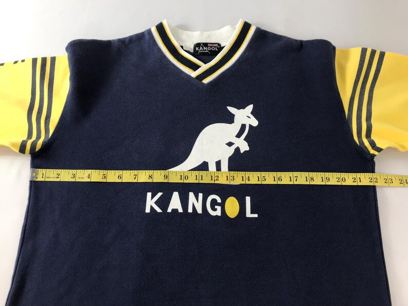 May include: Navy blue Kangol V-neck sweatshirt with yellow sleeves and black stripes. Features a white kangaroo logo and the brand name in white. The collar has white, black, and yellow trim. The sweatshirt measures approximately 24 inches across.
