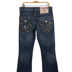 Könnte beinhalten: Blaue Jeans aus Denim mit Used-Look und einem True Religion-Markenpatch auf der Gesäßtasche. Die Jeans haben eine Bootcut-Form und ein regenbogenfarbenes Nähtdetail um die Gesäßtaschen.
