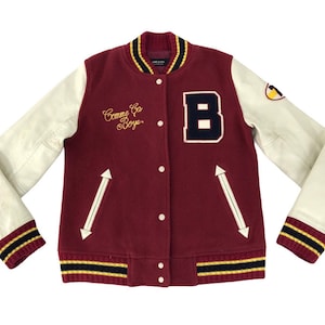 May include: Maroon varsity jacket with cream leather sleeves. Features a large navy blue "B" patch, gold script reading "Comme des Garcons Boys", and a yellow and black emblem on the sleeve.