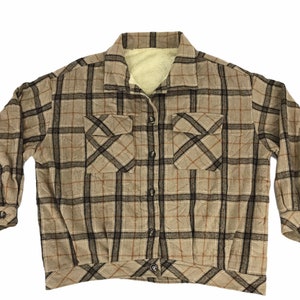 May include: A tan and black plaid shirt with a cream-colored interior lining. The shirt has two front pockets, long sleeves, and a button-down closure. The shirt is made of a soft, textured fabric.