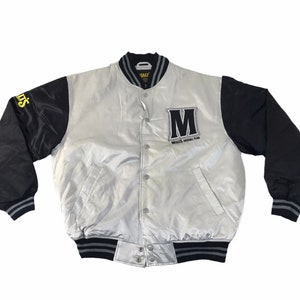 Könnte beinhalten: Silber-schwarze Baseballjacke mit dem Text "M" und "MALTS BASEBALL TEAM". Die Jacke hat schwarze Ärmel und einen silbernen Körper. Die Ärmel haben einen gelben Text auf dem linken Ärmel.