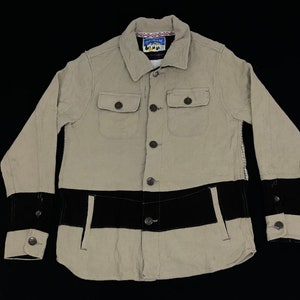 May include: A tan jacket with a black horizontal stripe across the midsection and black cuffs. The jacket has two chest pockets, a button-up front, and buttoned cuffs. A small patch with text is visible on the collar.