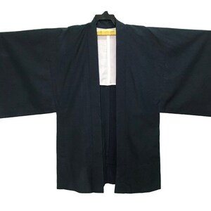 Puede incluir: Una bata estilo kimono azul marino oscuro con mangas anchas, colgada de una percha negra. La bata tiene un forro blanco visible en la abertura frontal. La prenda está hecha de una tela lisa, probablemente tejida.