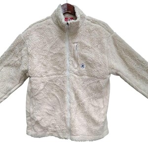 Può includere: Una giacca in pile color crema a maniche lunghe con collo alto e chiusura frontale a zip intera. Il capo presenta un esterno testurizzato e soffice, una tasca verticale con zip sul petto sinistro e una piccola toppa con logo circolare.