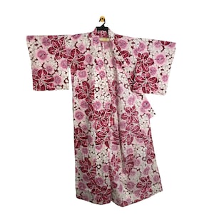 May include: A Japanese kimono, patterned with burgundy and white flowers, hangs on a black hanger. The garment's fabric is lightweight, with wide sleeves. The background is plain white. The kimono is a traditional garment.