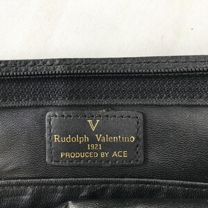 May include: Close-up of a black leather item with a zipper and a rectangular label. The label reads "Rudolph Valentino 1921 PRODUCED BY ACE" in gold lettering. The item appears to be a wallet or small bag.