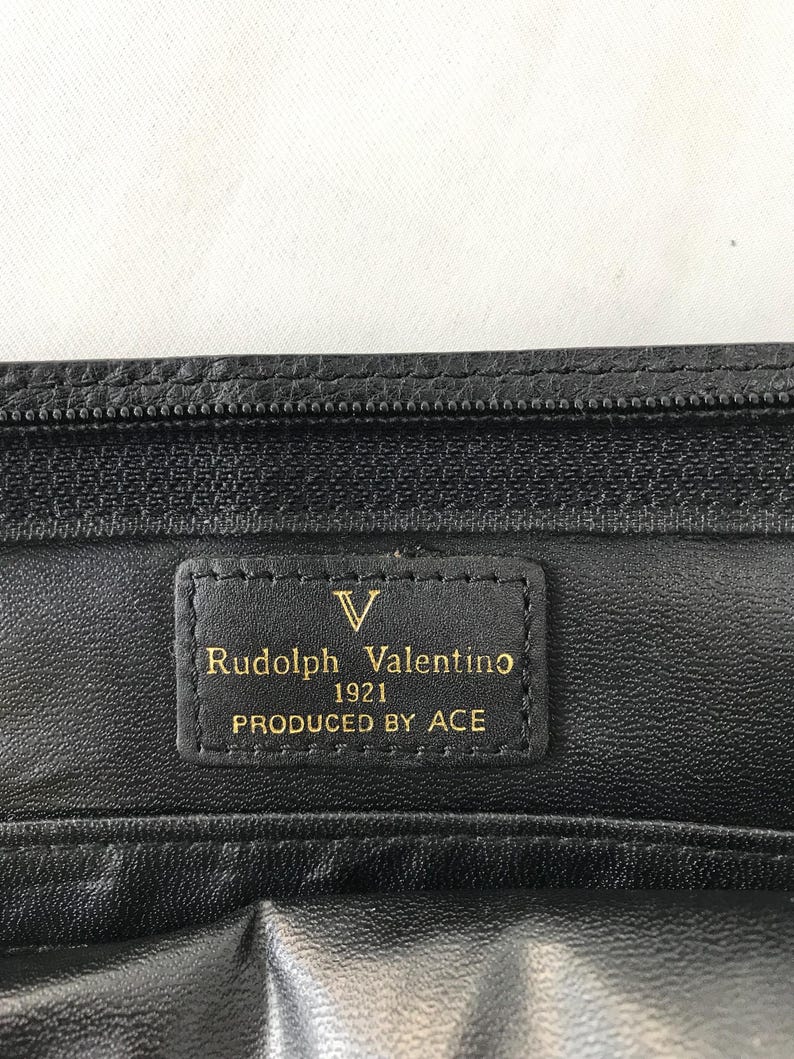 May include: Close-up of a black leather item with a zipper and a rectangular label. The label reads "Rudolph Valentino 1921 PRODUCED BY ACE" in gold lettering. The item appears to be a wallet or small bag.