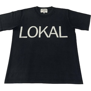 Puede incluir: Camiseta negra de manga corta con la palabra "LOKAL" impresa en letras blancas en la parte delantera. La camiseta tiene cuello redondo y un diseño sencillo y clásico, adecuada para uso casual.