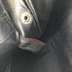 May include: Close-up of a black leather item with a silver snap closure. A small tag with red trim is visible, bearing text that reads "PRODUCED BY ... CHINA". The leather has a textured appearance.
