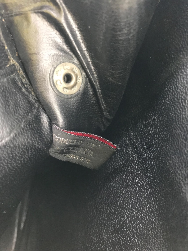 May include: Close-up of a black leather item with a silver snap closure. A small tag with red trim is visible, bearing text that reads "PRODUCED BY ... CHINA". The leather has a textured appearance.