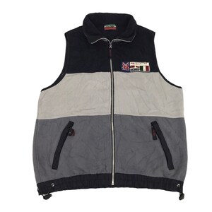 Könnte beinhalten: Eine farbblockierte Fleeceweste in Schwarz, Grau und Hellgrau. Die Weste hat einen durchgehenden Reißverschluss, einen Stehkragen und zwei Reißverschlusstaschen. Ein Aufnäher auf der Brust trägt die Aufschrift "Mally Grande Italia". Die Weste hat einen Kordelzug am unteren Saum.