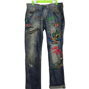 Op de afbeelding: Blauwe denim jeans met kleurrijke geborduurde ontwerpen, waaronder bloemen, vogels en de tekst "Nierba Mala Nunca Muere" en "Cerveza".
