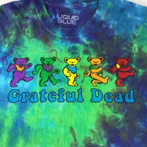 Puede incluir: Una camiseta tie-dye con el logotipo de la banda Grateful Dead con cinco osos bailando en colores arco&iacute;ris. La camiseta es azul, verde y morada con el texto "Liquid Blue" en la etiqueta.